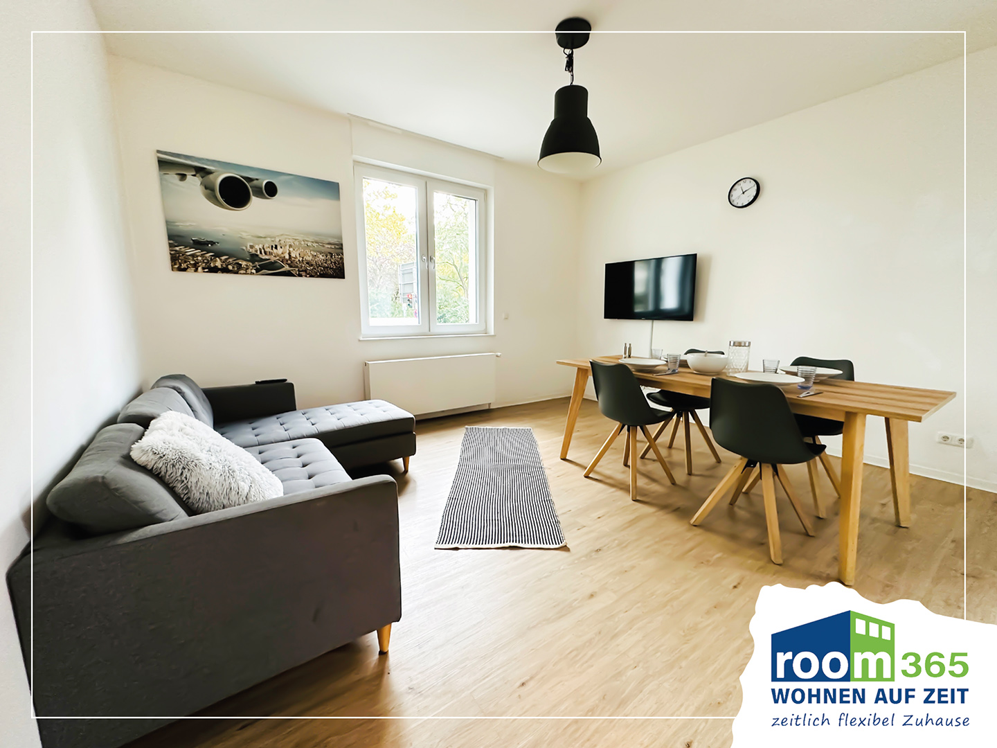 Das Wohnzimmer dieser Monteurwohnung 1 verfügt über ein graues Sofa, Esstisch mit Stühlen, Flachbild-TV, helle Holzböden und großes Fenster, gestreiften Teppich und schwarze Pendellampe.